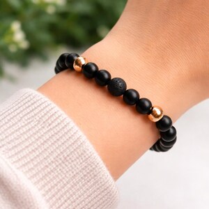 May include: Black beaded bracelet with gold-coloured accents and a lava stone bead. The bracelet is worn on a wrist, with a light-coloured sweater sleeve visible. The background is blurred with greenery.