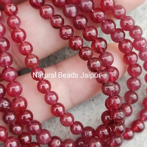 Könnte beinhalten: Nahaufnahme von mehreren Strängen kleiner, runder, tiefroter Perlen. Die Perlen sind aufgefädelt, und das Bild zeigt die Textur und Farbvariationen der Edelsteine. Der Text "Natural Beads Jaipur" ist im Bild zu sehen.