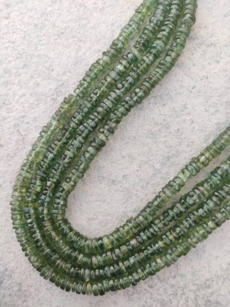 May include: A strand of green gemstone beads, with a faceted, slightly rounded shape. The beads are strung on a clear thread and are arranged in a loose, flowing pattern.