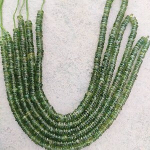 May include: A strand of green gemstone beads, likely peridot, with a faceted, round shape. The beads are strung on a thin thread and are arranged in a U-shape.
