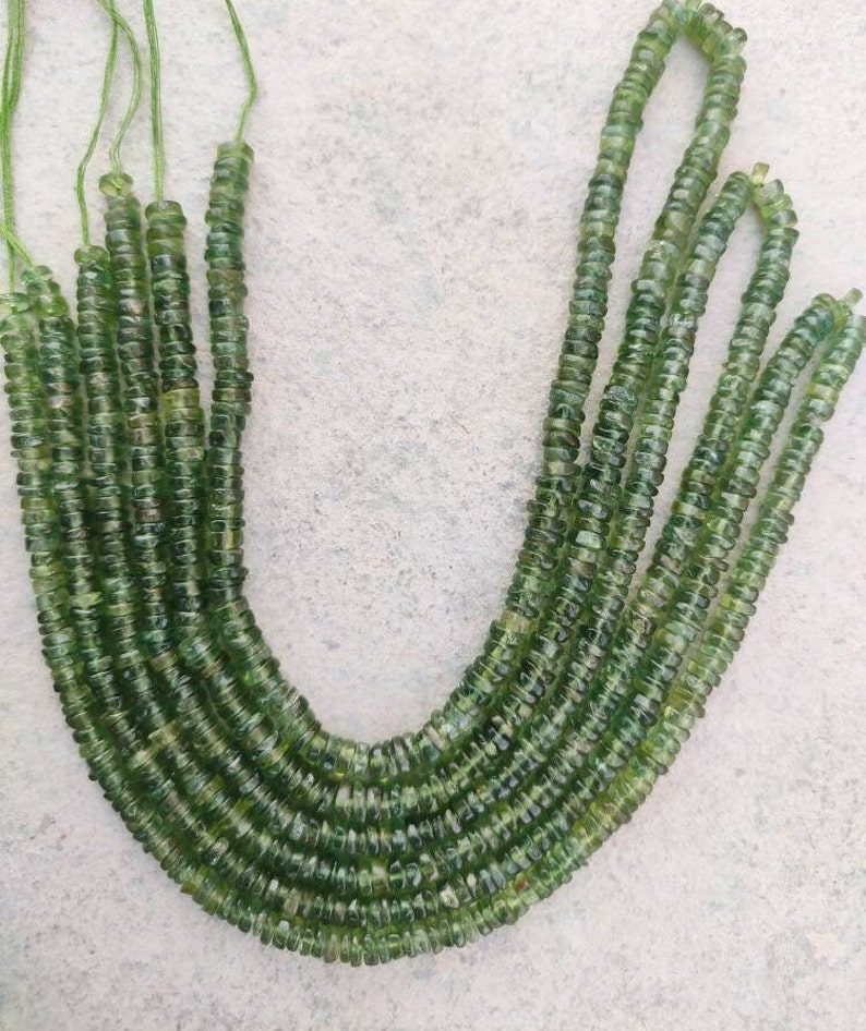 May include: A strand of green gemstone beads, likely peridot, with a faceted, round shape. The beads are strung on a thin thread and are arranged in a U-shape.