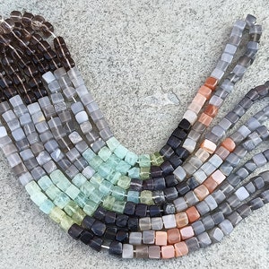 Puede incluir: Una colección de cuentas de piedras preciosas de forma cuadrada en varios colores, incluyendo marrón, gris, verde, negro y naranja. Las cuentas están dispuestas en un patrón de arcoíris.