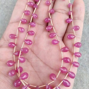 Puede incluir: Dos hilos de cuentas de piedras preciosas rosas. Cada cuenta tiene forma de lágrima y está ensartada en un alambre dorado. Las cuentas son de un color rosa vibrante y parecen pulidas.