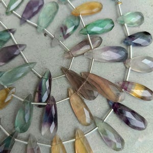 May include: Strands of faceted gemstone beads in various colors, including green, purple, yellow, and brown. The beads are teardrop-shaped and strung on silver-colored wire, with a white tassel in the background. Ideal for jewelry making.