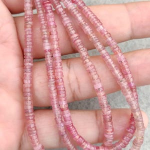 Puede incluir: Hebras de cuentas de piedras preciosas rosas. Las cuentas son pequeñas, planas y en forma de disco, ensartadas para formar collares. El color varía de rosa claro a oscuro, con ligeras variaciones de color. Las cuentas se muestran sobre un fondo neutro.