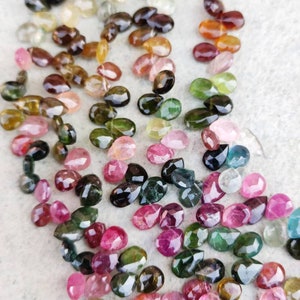 May include: A collection of colorful, faceted, teardrop-shaped gemstones. The gemstones are a mix of pink, green, brown, and black.