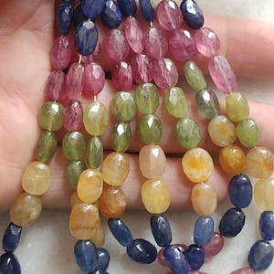 Puede incluir: Una colección de cuentas de piedras preciosas facetadas de forma ovalada ensartadas en múltiples hilos. Las cuentas muestran un degradado de colores que incluye azul profundo, rosa vibrante, verde oliva y amarillo cálido, en filas verticales.