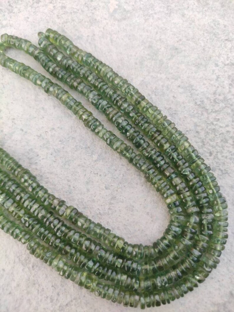 May include: A strand of green gemstone beads, with a faceted, round shape. The beads are strung on a clear thread and are arranged in a long, continuous line.