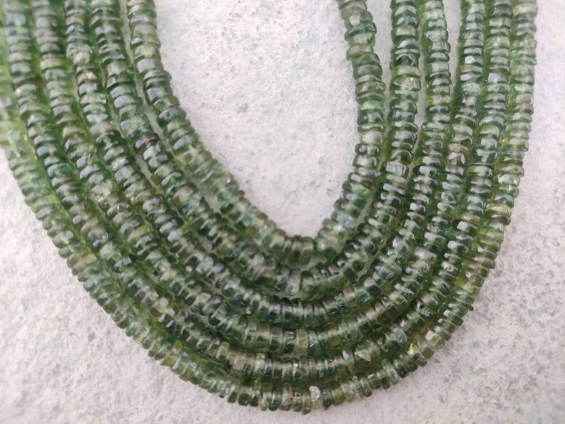 May include: A strand of green gemstone beads, each bead is faceted and has a slightly rounded shape. The beads are strung together to form a long necklace.