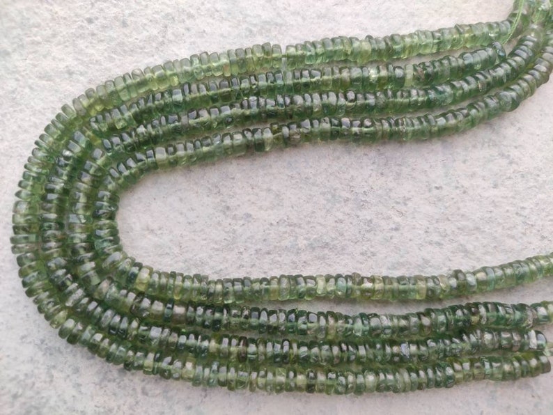 May include: A strand of green gemstone beads, each bead is faceted and has a slightly irregular shape. The beads are strung on a clear thread and are arranged in a loose, flowing pattern.
