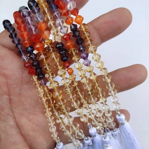 Op de afbeelding: Strengen gefacetteerde edelsteen kralen in diverse kleuren, waaronder oranje, paars, geel en helder. Elke streng is afgewerkt met een zilverkleurige dop en een lichtblauwe kwast. De kralen worden in een hand gehouden tegen een witte achtergrond.