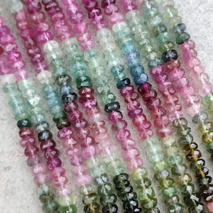 Op de afbeelding: Close-up van meerdere strengen toermalijn kralen. De kralen zijn gefacetteerd en vertonen een reeks kleuren, waaronder roze, groen, blauw en zwart. De kralen zijn aan elkaar geregen, waardoor een textuur ontstaat. Elke kraal is klein en rond.