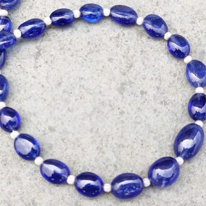 Op de afbeelding: Een ketting met ovale, diepblauwe kralen, afgewisseld met kleine witte kralen. De glanzende blauwe kralen zijn in een cirkelvormig patroon gerangschikt, wat een opvallend contrast in kleur en textuur creëert.