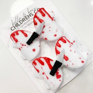 May include: Two white felt hair bows with red blood splatter and a black knife detail. The bows are on a white background with the text "belle & beau CHILDRENS CO.  bout & handmade hair - Las Vegas"