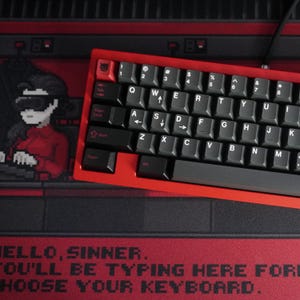 Può includere: Una tastiera meccanica rossa con tasti neri poggia su un tappetino da scrivania rosso e nero. La tastiera ha una cornice rossa e tasti neri con scritte bianche. Il tappetino presenta pixel art e il testo "HELLO, SINNER. YOU'LL BE TYPING HERE FOR... CHOOSE YOUR KEYBOARD."