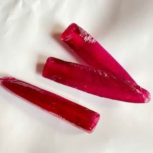 May include: Three long, pointed, ruby red crystals on a white surface.