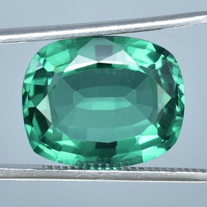 May include: A green gemstone with a cushion cut. The gemstone is held in place by a pair of tweezers.