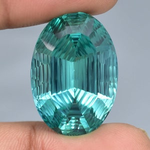 May include: A large, oval-shaped, faceted gemstone with a teal blue color. The gemstone has a complex, geometric cut with many facets.