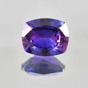 Könnte beinhalten: Ein kissenförmig geschliffener Edelstein in tiefem Lila, der das Licht durch seine Facetten reflektiert. Die polierte Oberfläche erzeugt eine spiegelähnliche Reflexion darunter. Die abgerundete quadratische Form zeigt die Klarheit und Farbe des Edelsteins.