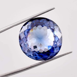 Puede incluir: Una piedra preciosa redonda y facetada de color azul con un centro azul claro. La piedra preciosa está sujeta con unas pinzas.