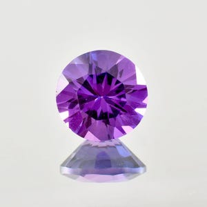 Puede incluir: Una gema púrpura redonda y facetada con talla brillante. La gema está colocada sobre una superficie reflectante, creando un efecto de espejo. Las facetas de la gema capturan y reflejan la luz, mostrando su color púrpura intenso.