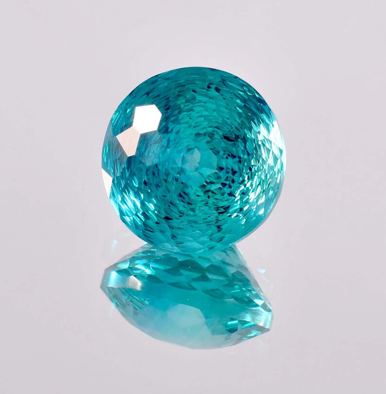 May include: A round, faceted gemstone with a teal blue color. The gemstone is reflecting light, creating a sparkling effect.