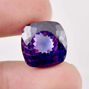 Könnte beinhalten: Ein facettierter, kissenförmiger Edelstein mit tiefvioletter Farbe, der im Zentrum in hellere Violett- und Blautöne übergeht. Der Edelstein wird zwischen zwei Fingern vor weißem Hintergrund gehalten, was seine Klarheit und Brillanz zeigt.