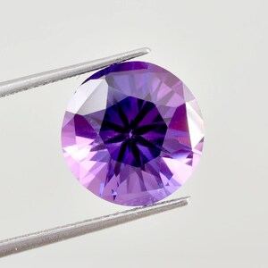 Peut inclure: Une pierre précieuse ronde et facettée, tenue par une pince argentée. La gemme présente un centre violet foncé qui s'estompe en nuances plus claires de lavande et de violet. La taille de la pierre crée un motif en étoile.