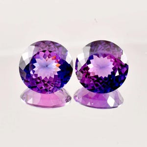 Puede incluir: Dos piedras preciosas redondas y facetadas con un degradado de morado a rosa. Las piedras preciosas están sobre un fondo blanco.