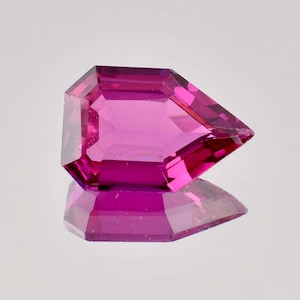 Puede incluir: Una gema facetada de color magenta con un corte geométrico único. Las facetas de la gema reflejan la luz, creando una exhibición brillante. La piedra se encuentra sobre una superficie reflectante, realzando su atractivo visual. Un ejemplo de joyería fina.