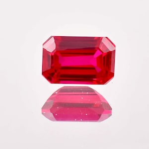 May include: A vibrant red gemstone with a rectangular emerald cut. The gemstone is faceted and has a high polish.