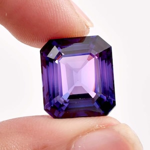 May include: A rectangular-cut gemstone in shades of purple, held between fingers. The gem's facets are clearly visible, reflecting light. The colour shifts from a dark purple at the edges to a lighter, pinkish tone in the centre.