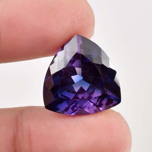 Peut inclure: Une pierre précieuse triangulaire de couleur violet foncé avec une taille facetée. La pierre précieuse est tenue dans une main.