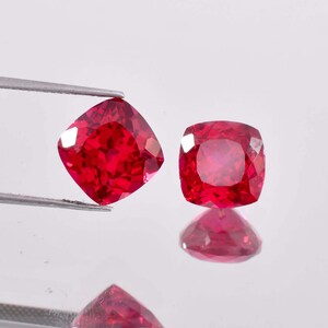 Puede incluir: Dos piedras preciosas de talla cojín roja, probablemente rubíes, se muestran sobre una superficie blanca. Las piedras preciosas están sujetas por unas pinzas.