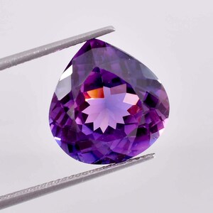 Puede incluir: Una piedra preciosa de amatista en forma de pera con una inclusión en forma de estrella en el centro. La piedra preciosa es de color morado oscuro con toques de rojo y naranja.