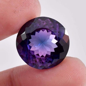 Puede incluir: Una piedra preciosa de amatista redonda y facetada con un degradado de color púrpura y azul. La piedra preciosa tiene un patrón de estrella en el centro.