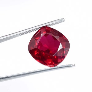 May include: A deep red cushion-cut gemstone held by tweezers. The gemstone is faceted and has a high shine.