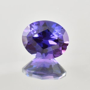 Könnte beinhalten: Ein facettierter, oval geschliffener Edelstein mit tiefvioletter Farbe. Der Edelstein ist hochreflektierend, mit mehreren Facetten, die das Licht einfangen und eine brillante Darstellung erzeugen. Der Edelstein ist vor einem hellen Hintergrund platziert, mit seiner Reflexion darunter sichtbar.