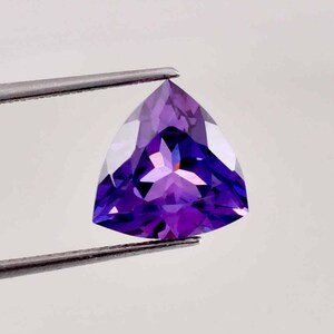 Puede incluir: Una piedra preciosa de amatista tallada en forma triangular con un tono púrpura. La piedra preciosa está sujeta por unas pinzas sobre un fondo blanco.