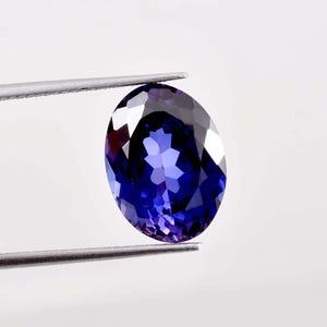 May include: A deep blue, oval-cut gemstone held by tweezers. The gem's facets reflect light, creating a sparkling effect. The stone's colour ranges from dark blue to lighter shades, with hints of purple. The tweezers are silver-coloured, and the background is plain white.