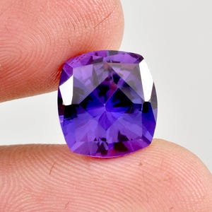 Könnte beinhalten: Ein facettierter, kissenförmiger Edelstein in tiefem Lila. Die Oberfläche des Edelsteins reflektiert das Licht und zeigt seine komplizierte innere Struktur. Der Stein wird zwischen zwei Fingern gehalten, was seine Größe und Form hervorhebt. Für Schmuck.