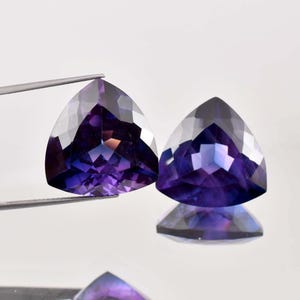 Puede incluir: Dos piedras preciosas triangulares facetadas con un degradado de púrpura a transparente. Las piedras preciosas están talladas en forma triangular y tienen un alto nivel de brillo.