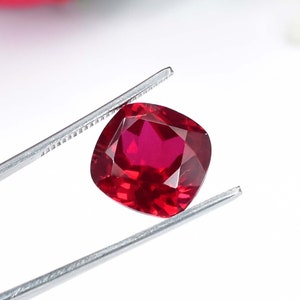 Pode incluir: Uma gema de corte almofada de cor vermelha profunda, segurada por pinças. A gema é facetada e tem um brilho brilhante.