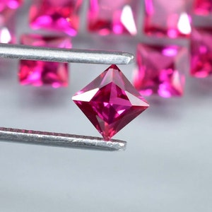 May include: A vibrant pink square-shaped gemstone, held by tweezers, with a faceted cut and a high-gloss finish. The gemstone is a ruby, a precious stone known for its deep red color.