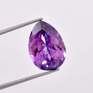 May include: A pear-shaped amethyst gemstone with a purple hue, held by tweezers against a white background.