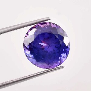 Puede incluir: Una piedra preciosa redonda y facetada con un degradado de color de púrpura a azul. La piedra preciosa está sujeta por unas pinzas.