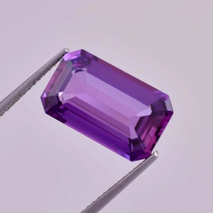 以下が含まれることがあります： エメラルドカットの濃い紫色のアメジストの宝石。宝石はピンセットで保持されています。