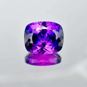 Könnte beinhalten: Ein kissenförmiger Edelstein in leuchtendem Lila, der das Licht mit faszinierender Brillanz reflektiert. Die Facetten des Steins erzeugen ein Spiel von Licht und Schatten, das seine Tiefe und Schönheit unterstreicht. Der Stein liegt auf einer reflektierenden Oberfläche.