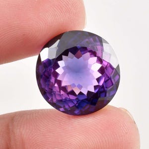 Puede incluir: Una piedra preciosa de amatista redonda y facetada con un degradado de color de púrpura a púrpura azulado. La piedra preciosa está tallada en un patrón de estrella.