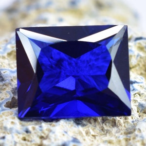 Könnte beinhalten: Ein tiefblauer Edelstein mit quadratischer Form und facettenreichem Schliff. Der Edelstein ist auf einer rauen, strukturierten Oberfläche platziert.